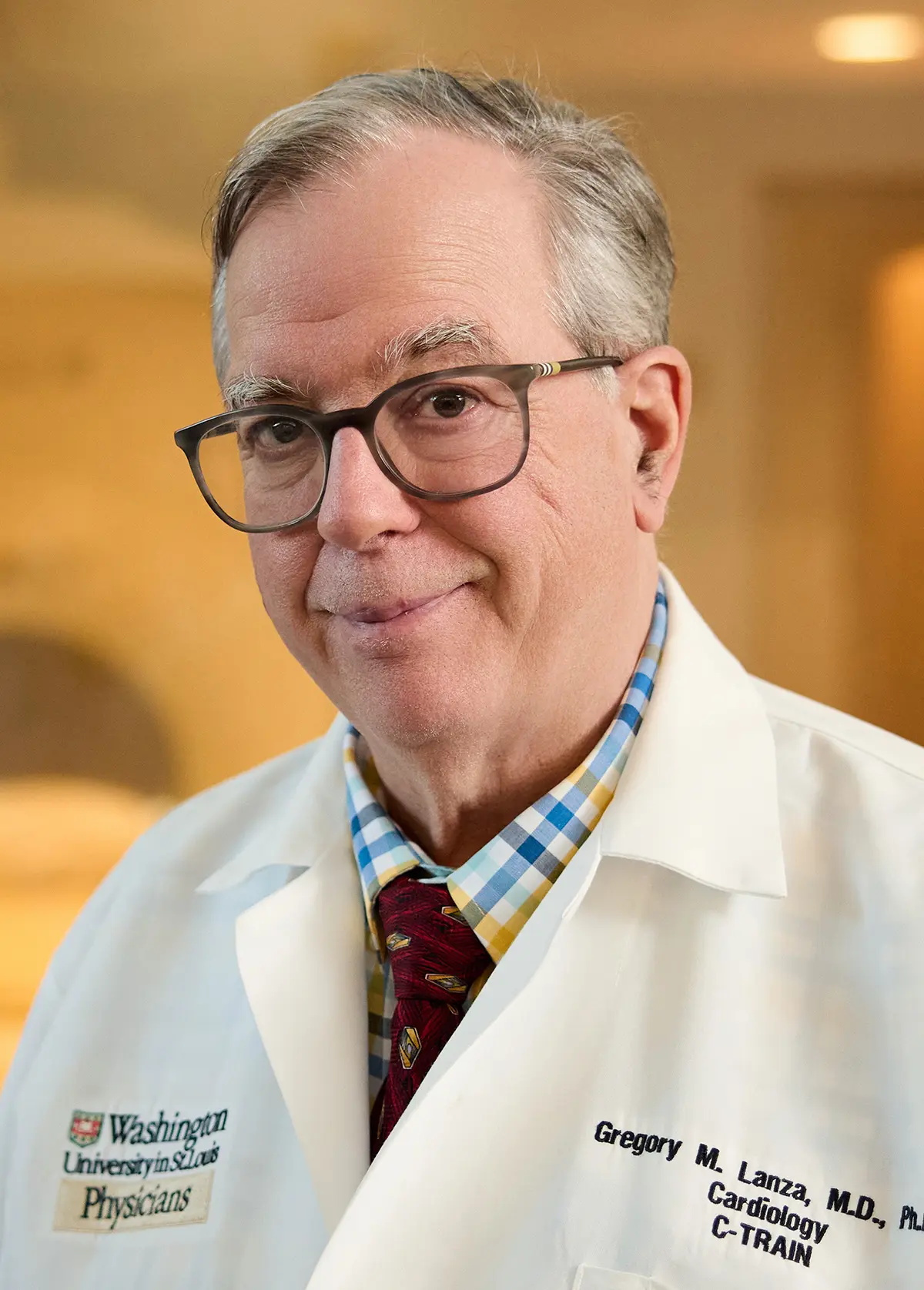 Portrait orientation photograph headshot of Gregory M. Lanza, M.D., Ph.D., an older elderly stage physician with gray hair and eyeglasses as he is grinning while wearing a white lab coat over a blue and yellow checkered button-up dress shirt and a patterned tie; The Washington University in St. Louis Physicians logo is visible on his coat