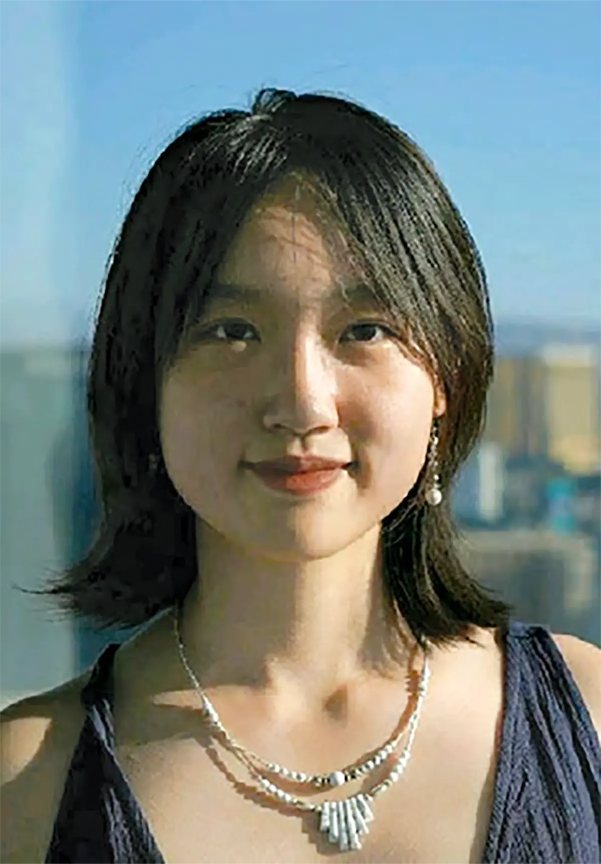 Portrait orientation photograph headshot of Heidi Minghao He, a young woman grinning with short dark black hair, wearing a dark blue sleeveless dress top and a layered pearl and silver necklace, against a blurred background of a bright sky and city buildings; She is also wearing dangling pearl earrings