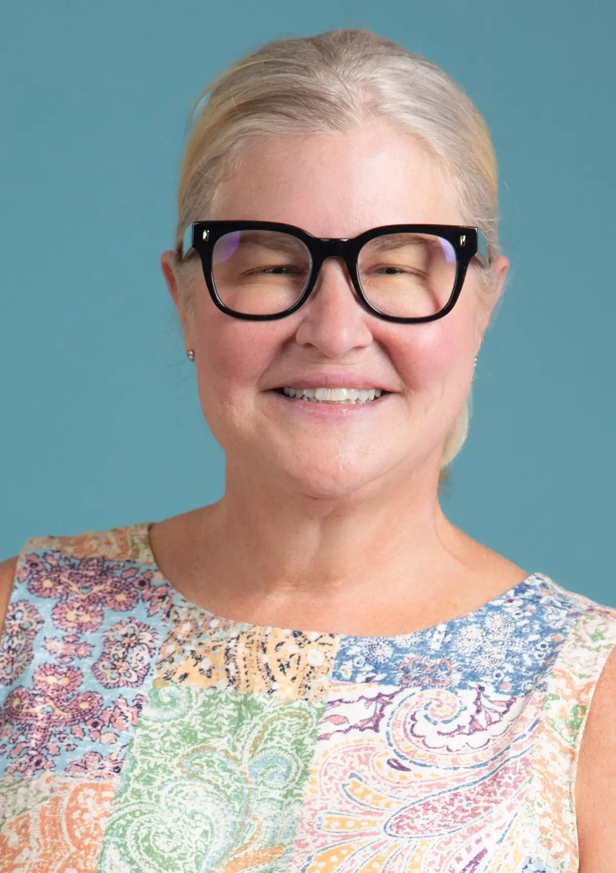 Portrait orientation photograph headshot of Jen Millard, who is an older elderly stage woman smiling with short blonde/graying hair pulled back, wearing black-framed eyeglasses and a colorful patterned blouse top, against a solid teal background