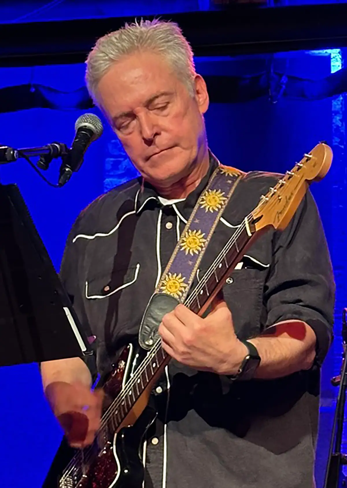 Portrait orientation photograph of Jonathan Salem Baskin, an older man with graying hair, wearing a black button-up dress shirt with white piping and a guitar strap with sun and star designs, playing an electric guitar on a stage with blue lighting behind him and a microphone stand nearby