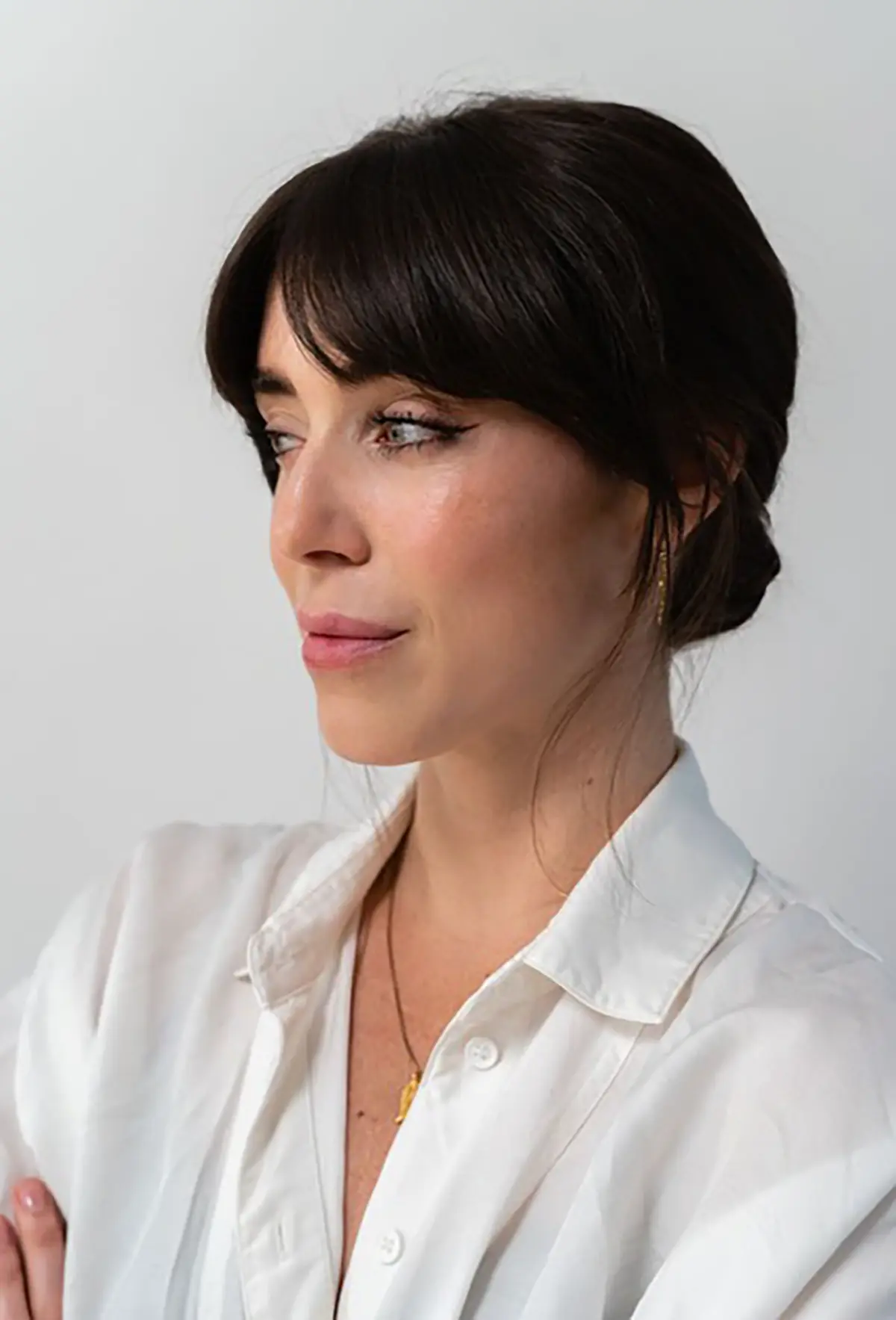 Portrait orientation photograph headshot of Melyn McKay, a young woman with dark black hair styled with bangs and a low bun, wearing a white collared button-up dress shirt and a delicate necklace, as she is looking slightly away from the camera against a soft white background