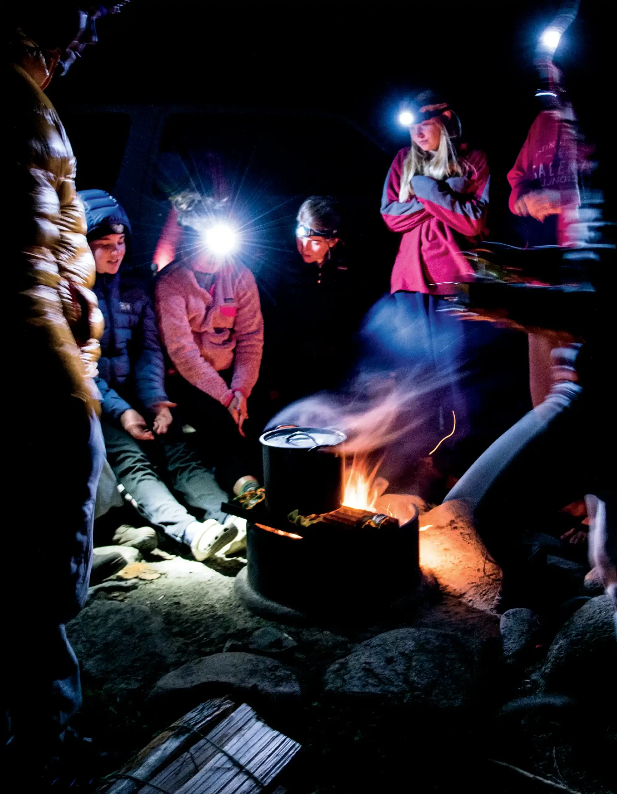 group of students gathered around fire while wearing hiking gear including thick coats and headlamps