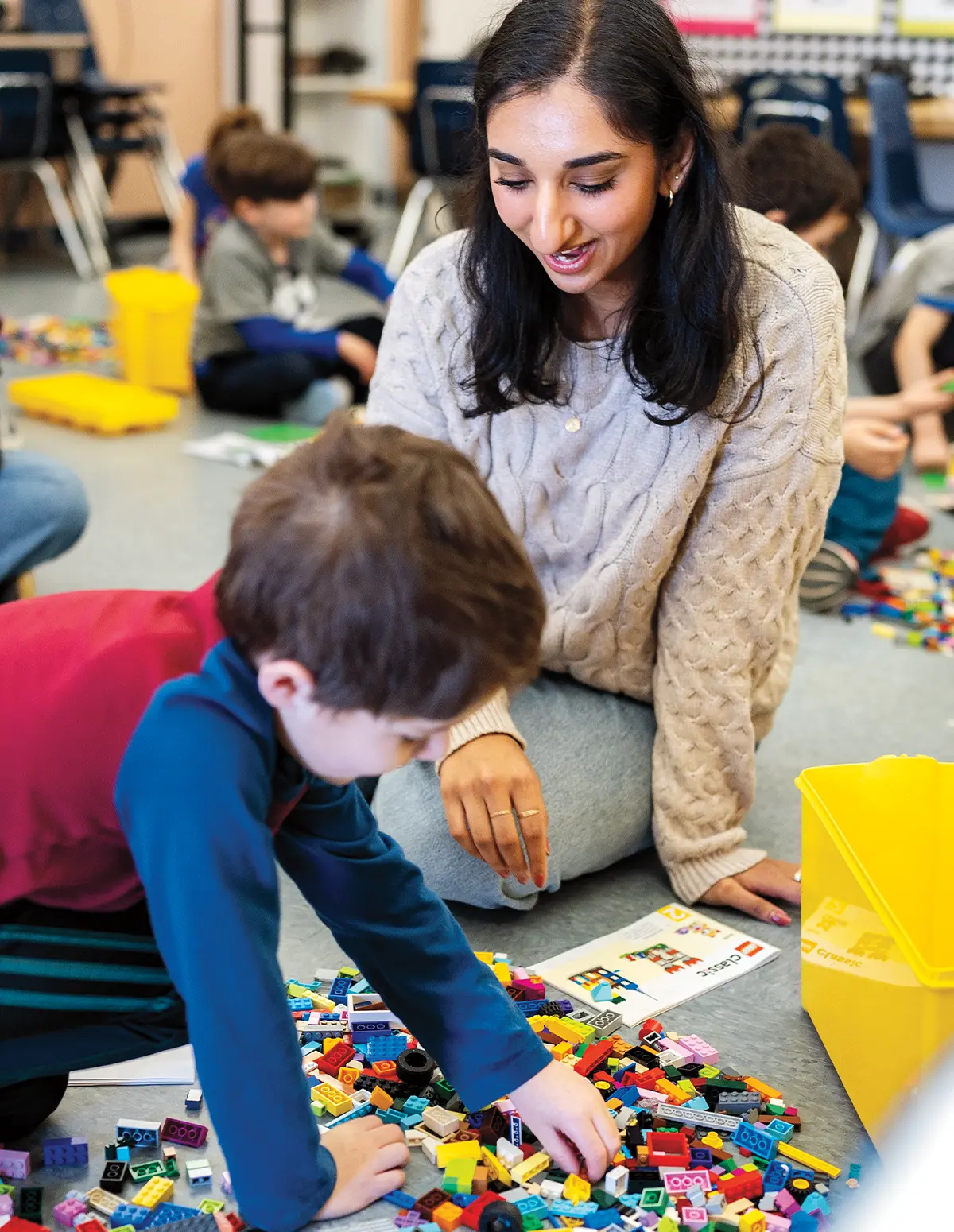 Ishani Wakhlu sits on the floor of a classroom and smiles as she watches a young boy play with legos