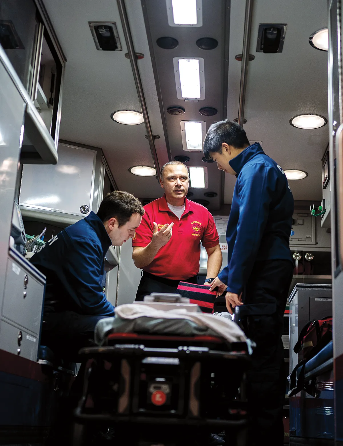 Department deputy EMS chief Everett Flannery wears a red embroidered EMS Chief polo while showing two uniformed Colby students the various items used in the cabin of an emergency service vehicle