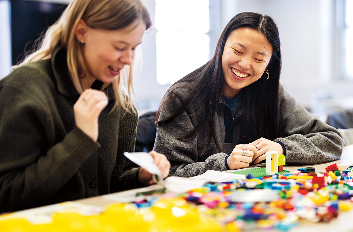 two young woman smile as they sit at a table in front of a large colorful pile of small legos