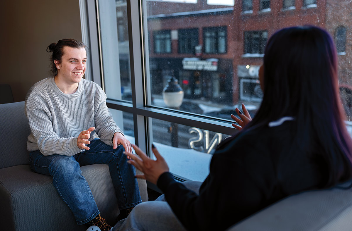 Matt Matheny smiles as they sit in an armchair across from a peer also seated in, both egaging beside a large window overlooking a wintry street