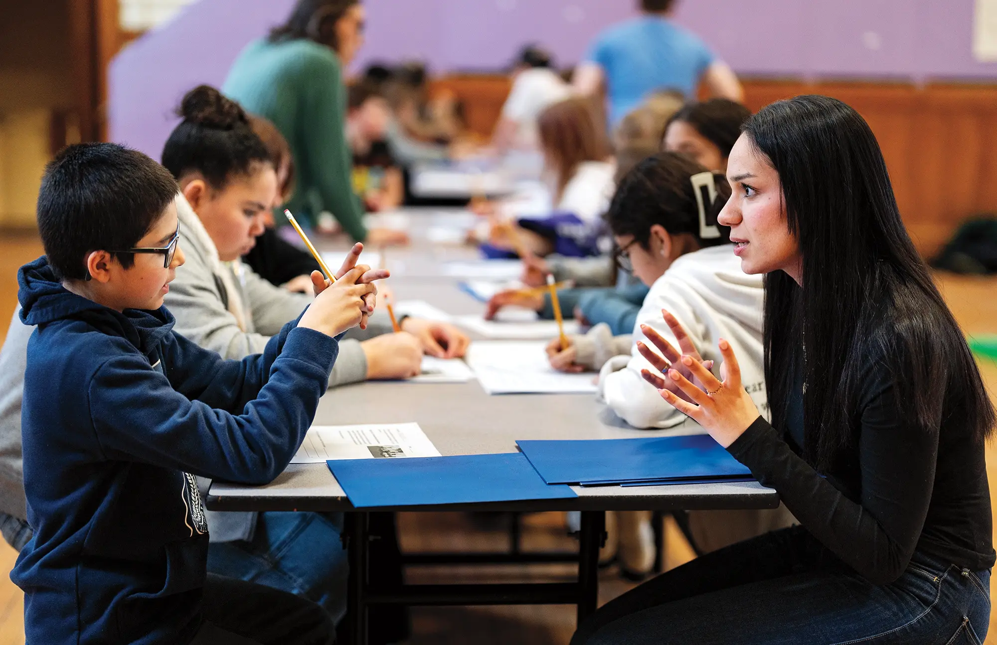 A tutoring setting with a group of students sitting at tables, engaged in a learning activities. Crystie Frometa, a Colby student is interacting with one of the students in the foreground.