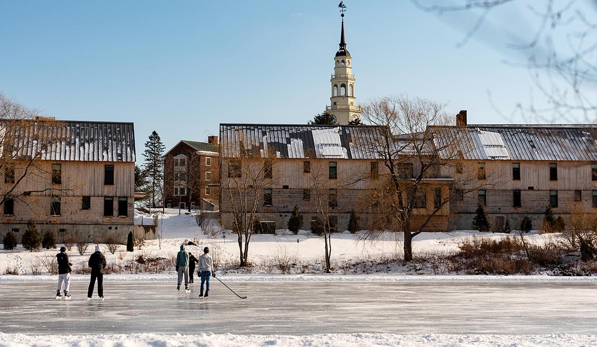 Outdoor photograph capturing a winter scene on the Colby College campus during the day where a group of students are ice skating and playing pond hockey