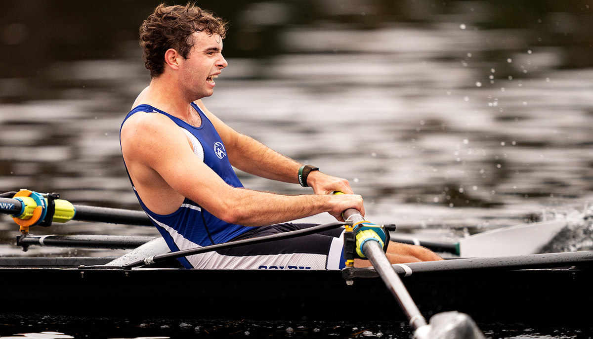 A close-up action photograph capturing a male Colby College rower competing on a river or lake with an intense physical exertion expression
