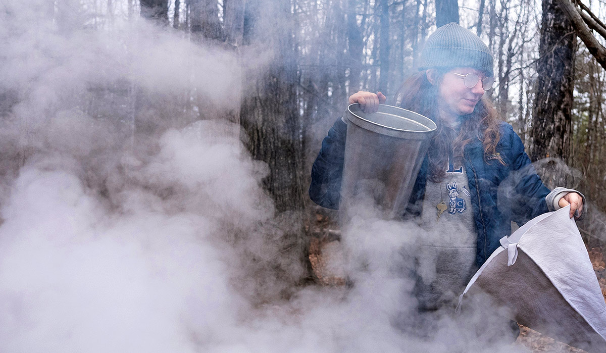 A medium, eye-level outdoor photograph capturing a student working amidst a thick cloud of white steam or smoke in a wooded area
