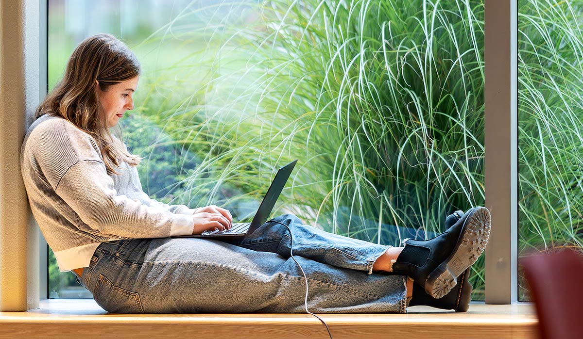 A horizontal, full-length indoor photograph capturing a female student studying comfortably on a wide wooden windowsill