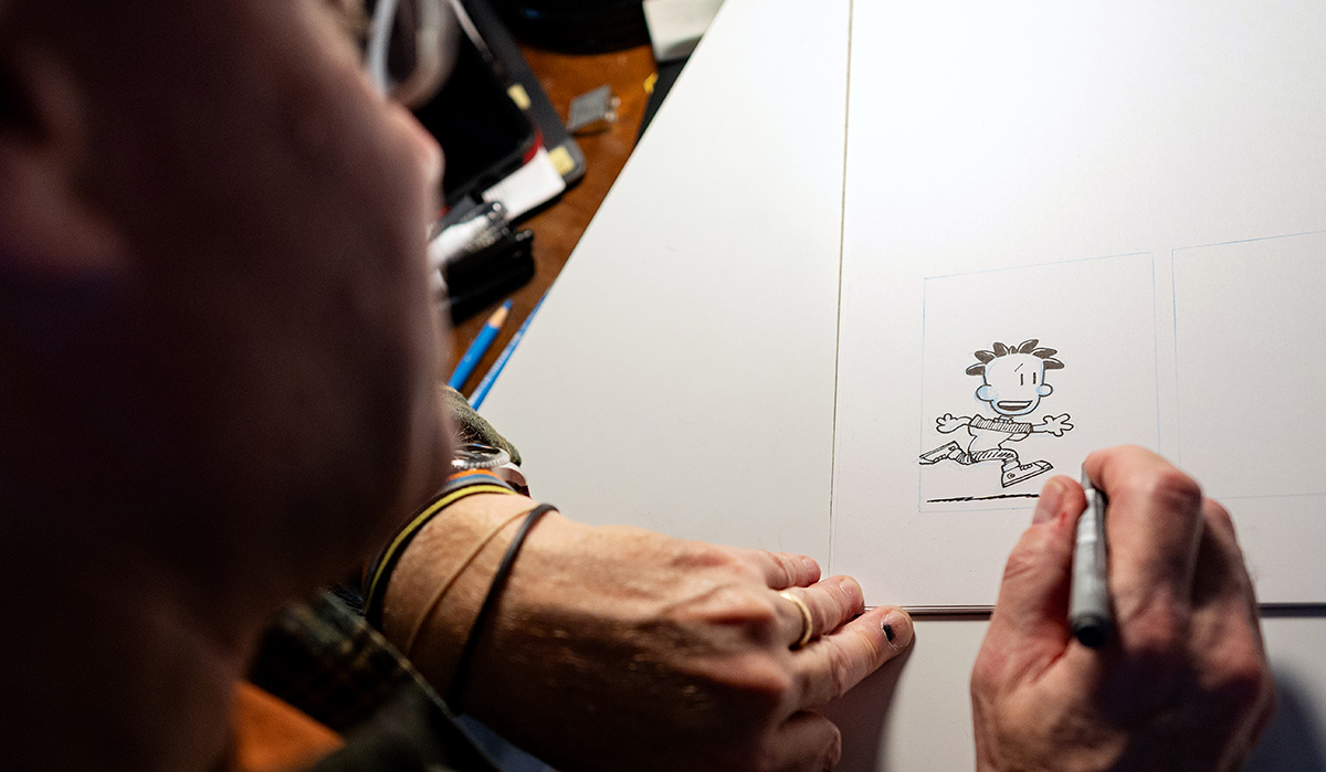 An over-the-shoulder, high-angle close-up photograph capturing an artist drawing a cartoon strip on a white board or thick paper