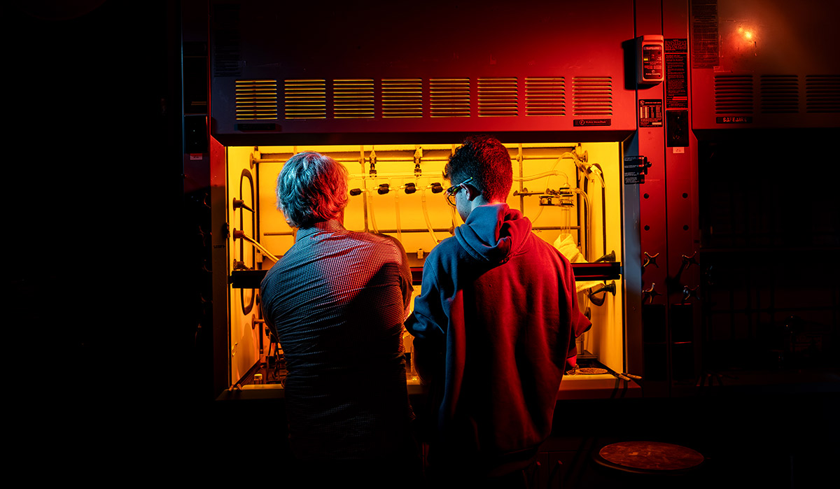 A wide, dramatic low-light photograph capturing a professor and a student from behind as they work together at a chemical fume hood