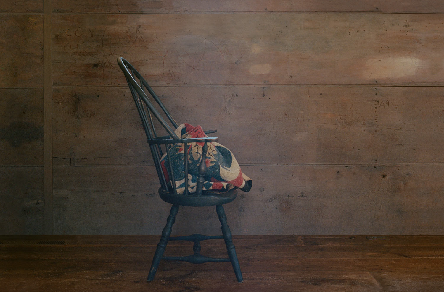 A dark green wooden spindle-back chair holding a colorful patterned blanket, set against a rustic, weathered wood-plank wall.