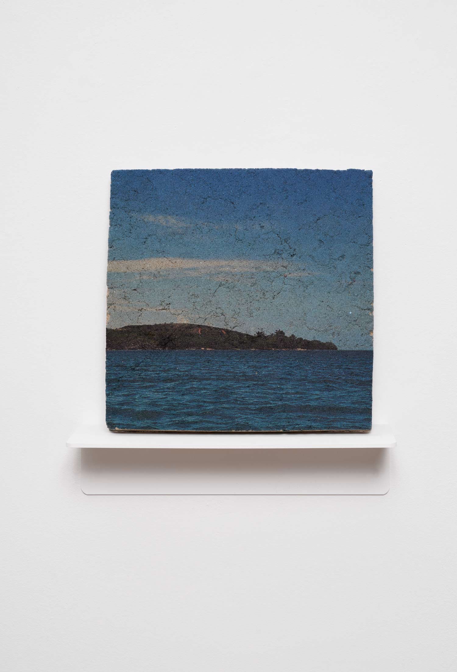 A square, textured artwork depicting a dark island in a blue sea under a twilight sky, displayed on a small white gallery shelf.