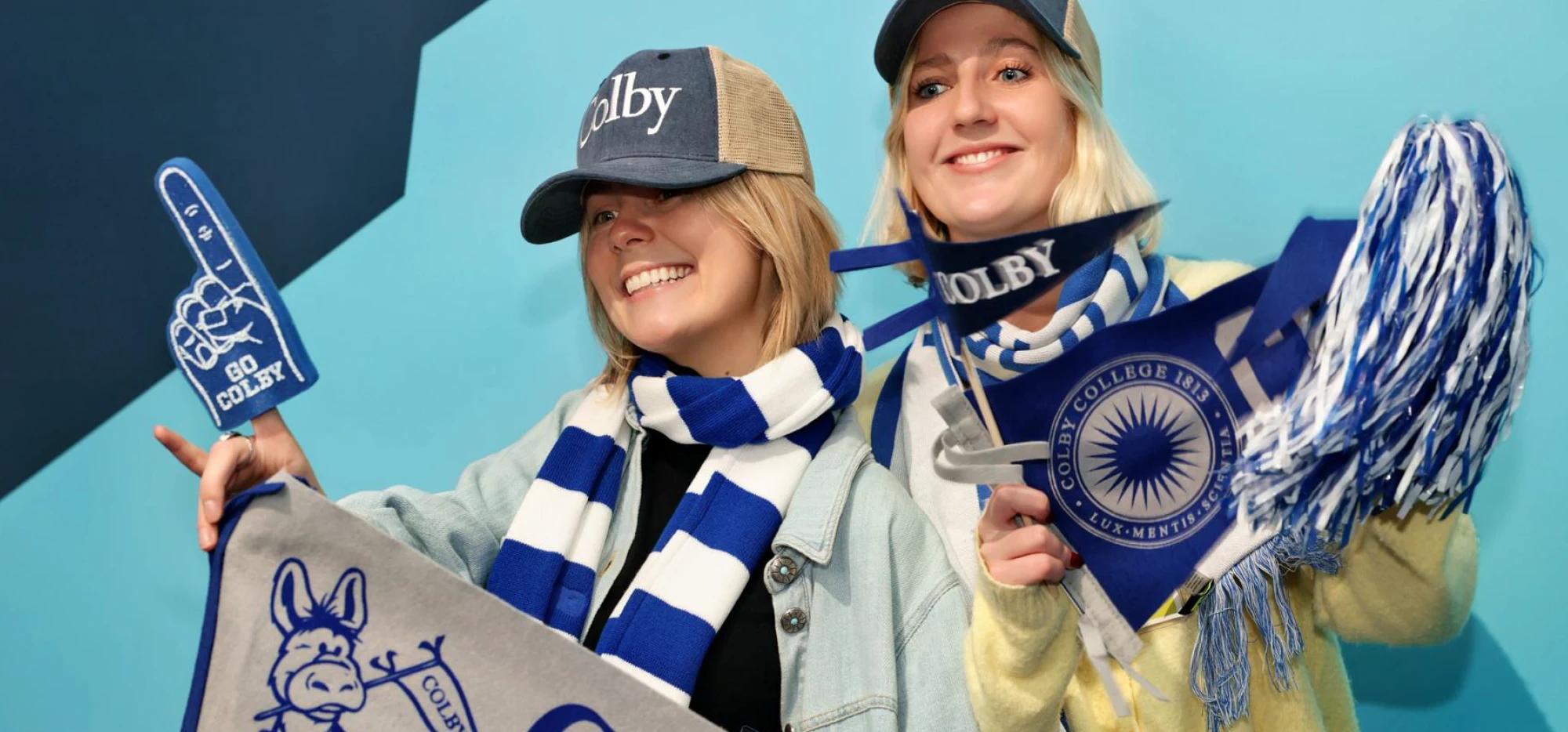 Two smiling students wearing Colby College hats and scarves wave a blue-and-white foam finger and pennants.