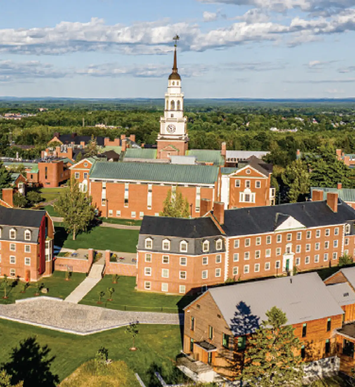 An elevated view of a college campus featuring red brick buildings, a prominent white clock tower steeple, and green lawns, with a vast forest extending into the background under a partly cloudy sky.