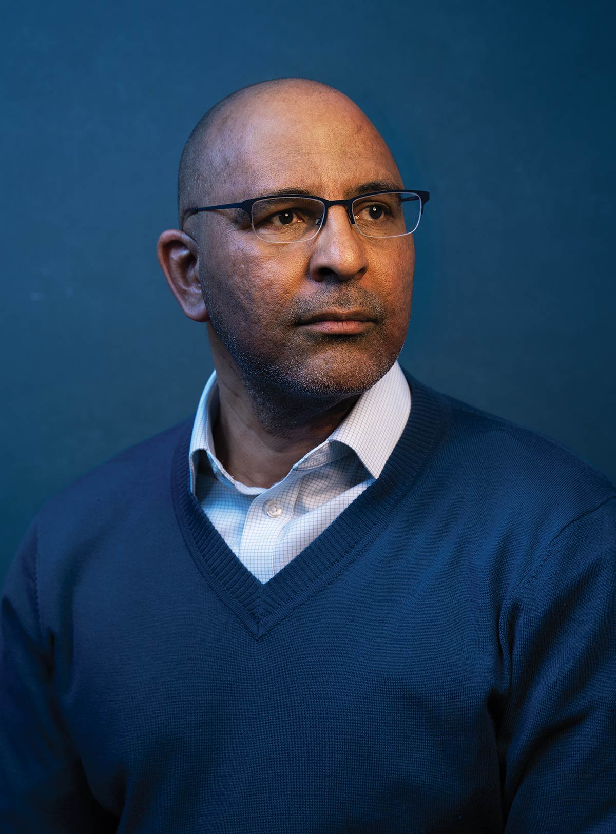 A bald man wearing glasses, a light checkered shirt, and a blue V-neck sweater poses looking thoughtfully to the side against a solid dark blue background.