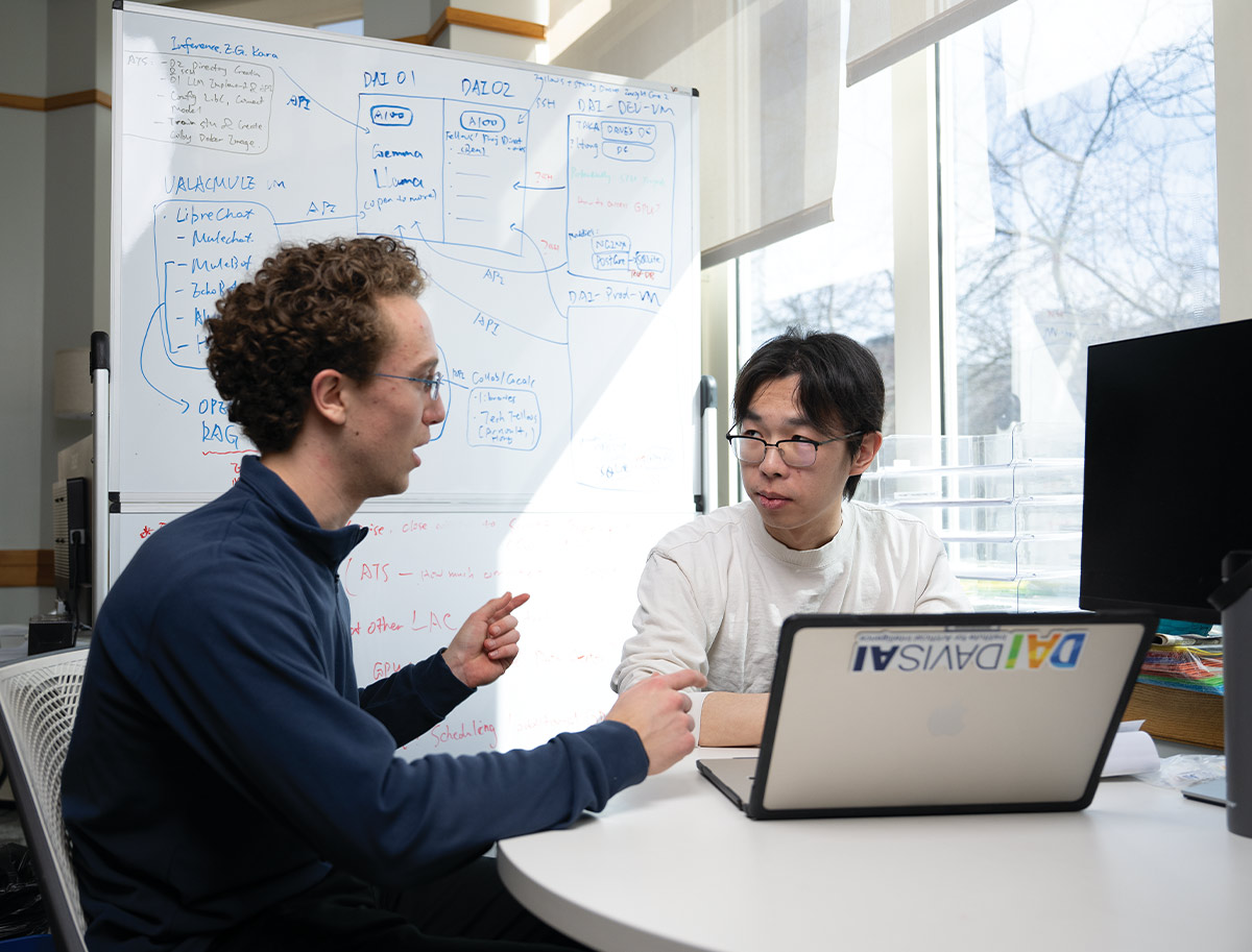 Two men sit at a desk collaborating in front of an open laptop. One gestures while speaking to the other, who listens intently. A large whiteboard covered in technical notes and diagrams stands in the background.