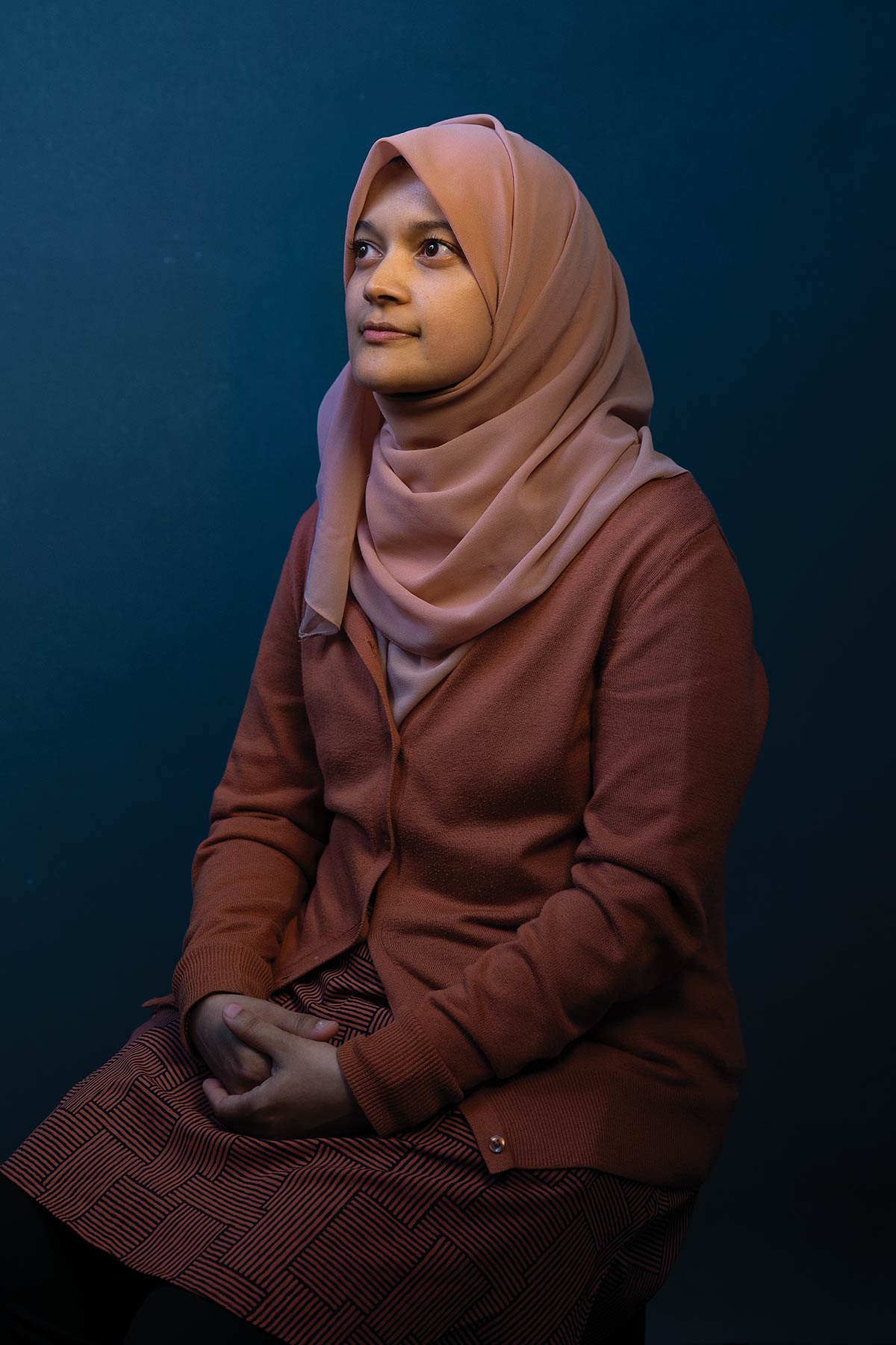 A woman wearing a pink hijab and a terracotta cardigan sits with her hands clasped in her lap, looking thoughtfully upward against a solid dark teal background.