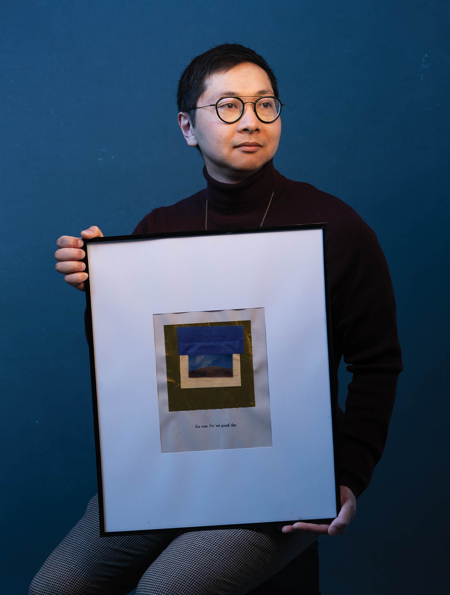 A man wearing glasses and a maroon turtleneck holds a framed piece of artwork featuring text that reads "this was the last good day." He poses looking to the side against a solid dark blue background.