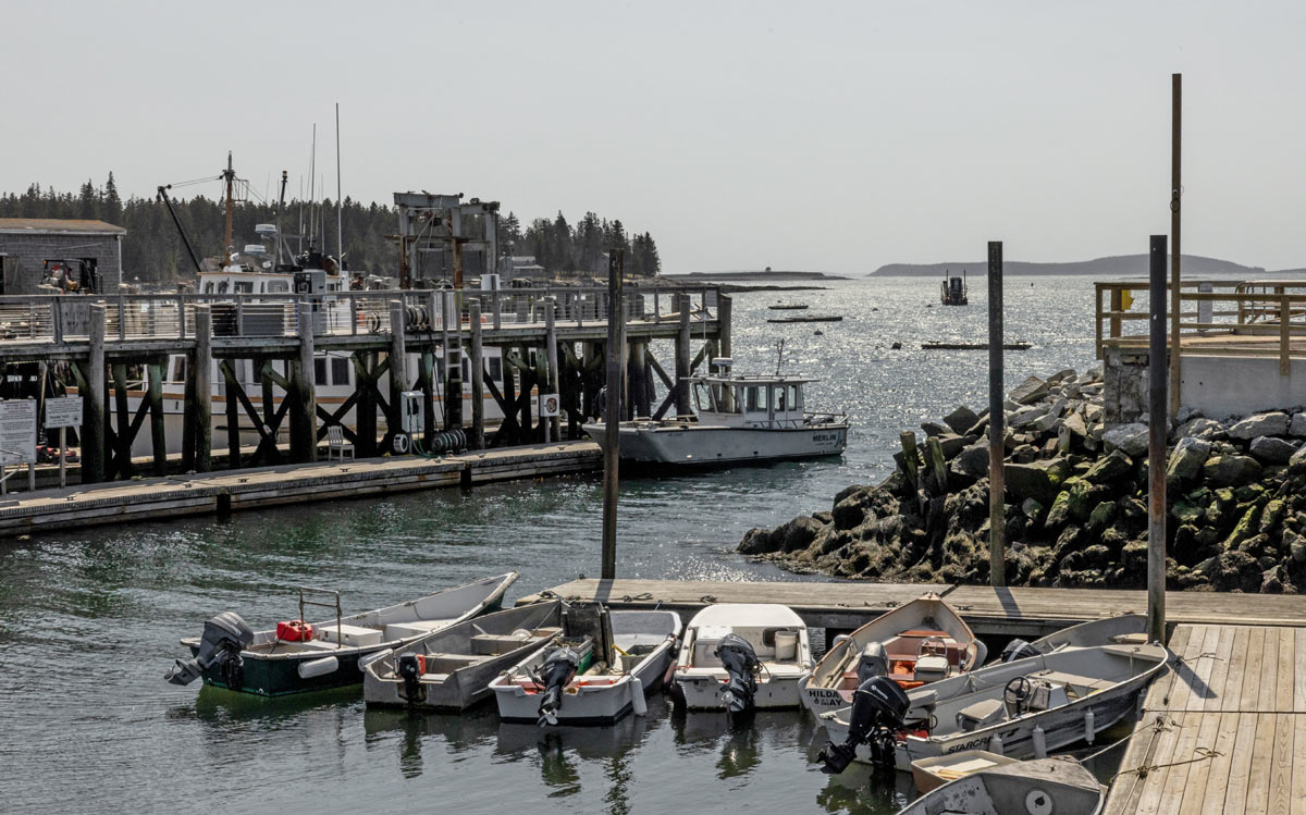 Several small motorboats are tied up at a wooden pier in a coastal harbor, with a larger boat and structures visible under a hazy sky.