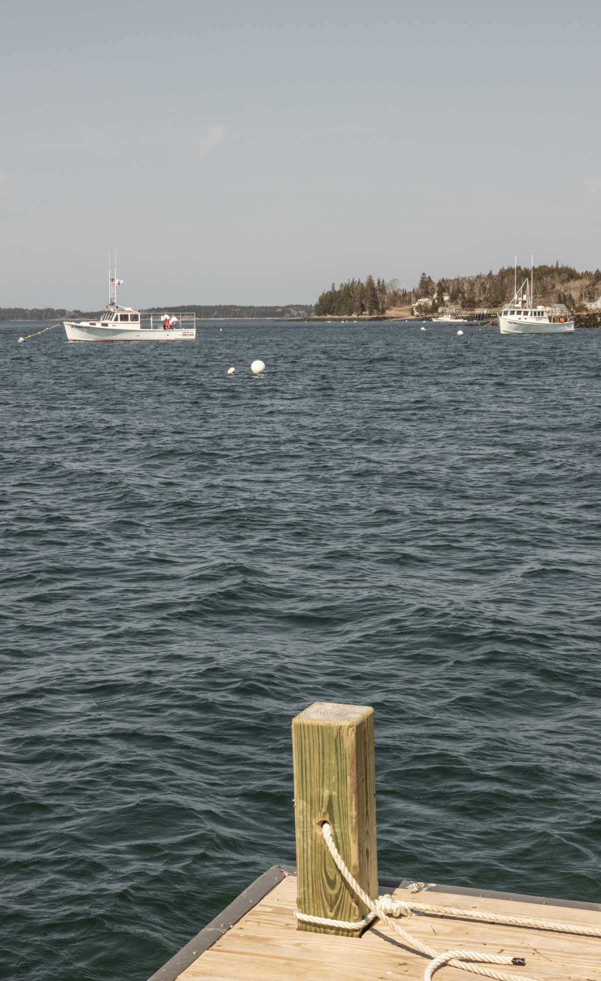 White fishing boats float on a dark, choppy sea under a grey sky, viewed from a wooden dock with a thick mooring post in the foreground.