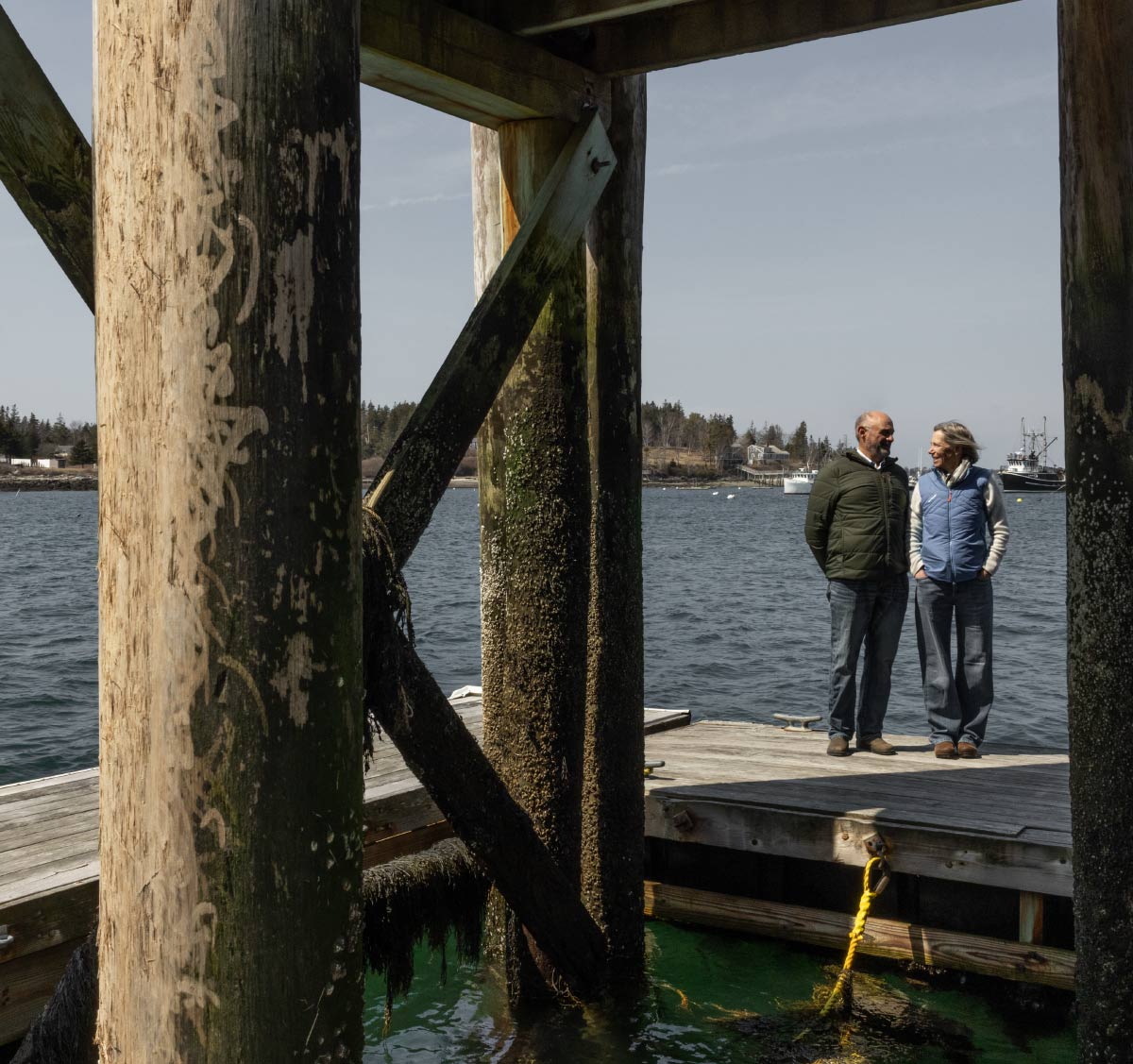 A man and a woman stand talking on a wooden dock next to large, textured wooden support pillars over dark ocean water.