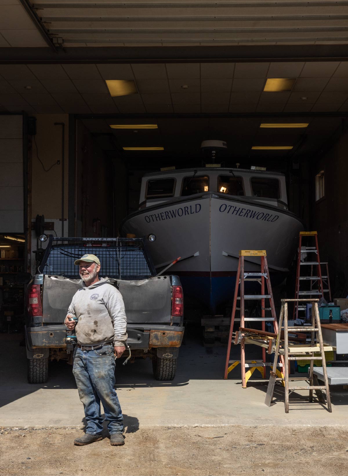 A worker in a sweatshirt and cap stands in a workshop with ladders and a pickup truck, in front of a large white boat named "OTHERWORLD".