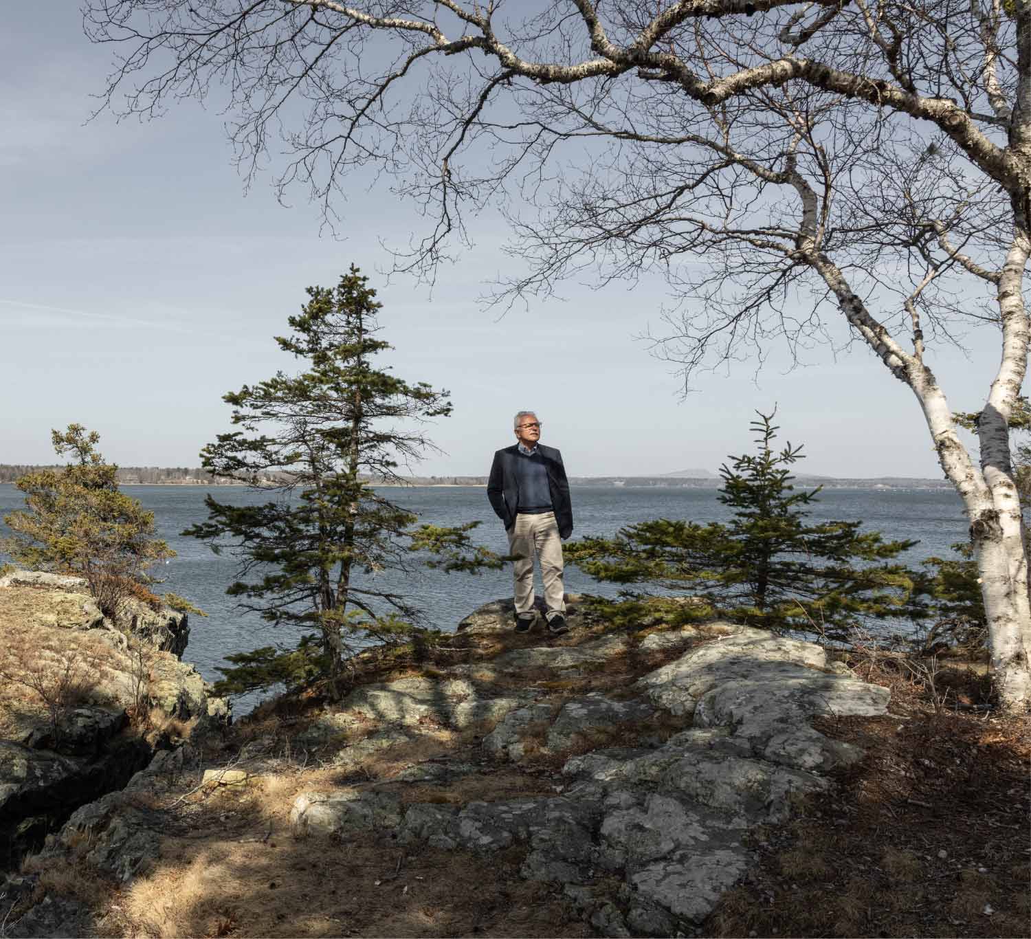 A man stands on a rocky, tree-lined point overlooking a wide body of water on a clear day, framed by bare birch branches.