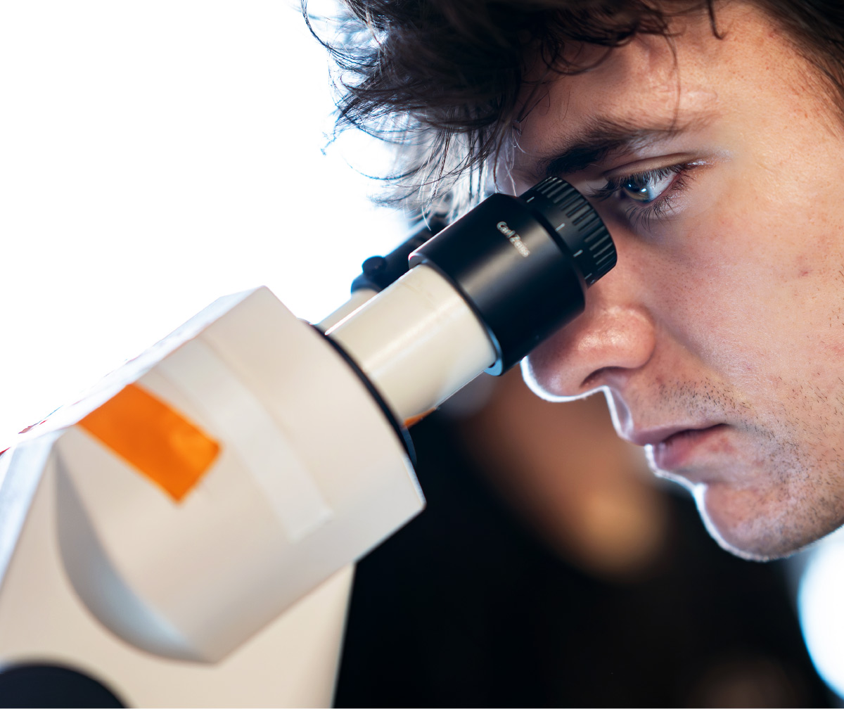 A close-up profile view of a young scientist looking intently through the eyepiece of a high-powered laboratory microscope.