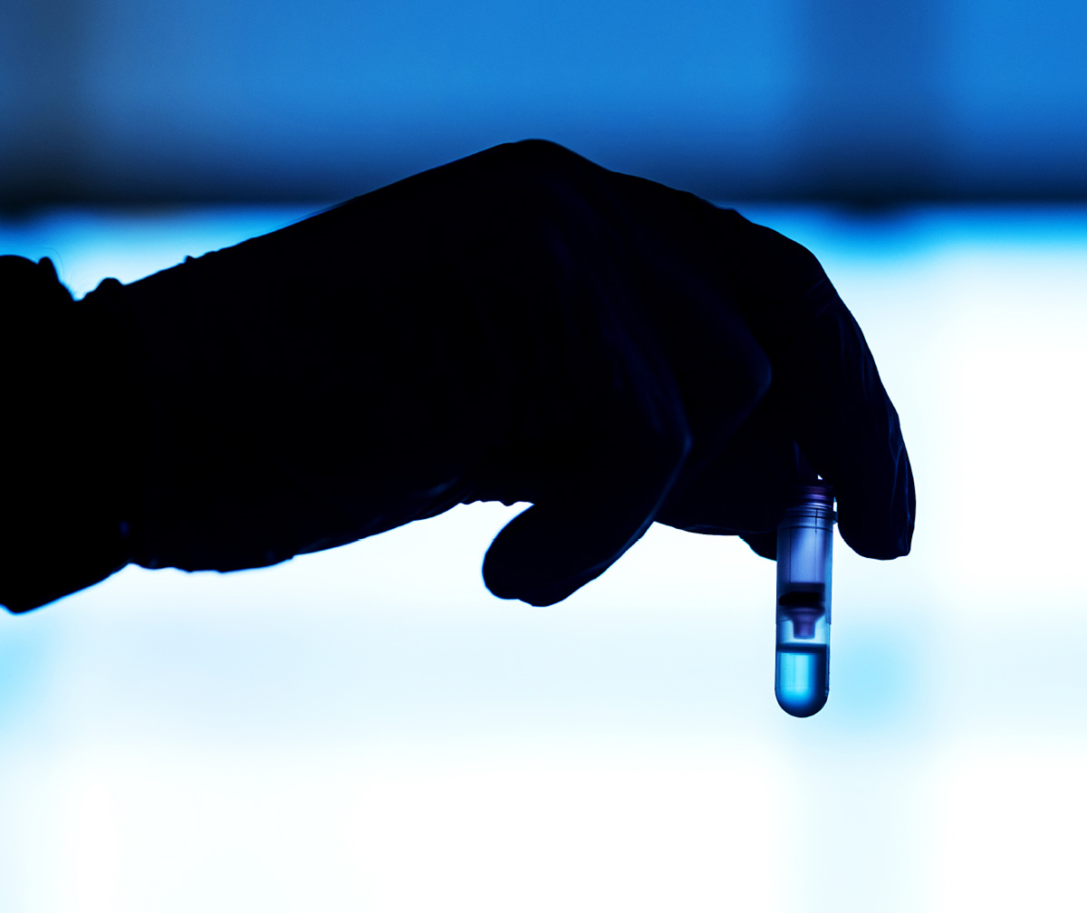 A stark silhouette of a gloved hand holding a small plastic vial filled with clear liquid against a glowing, bright blue background.
