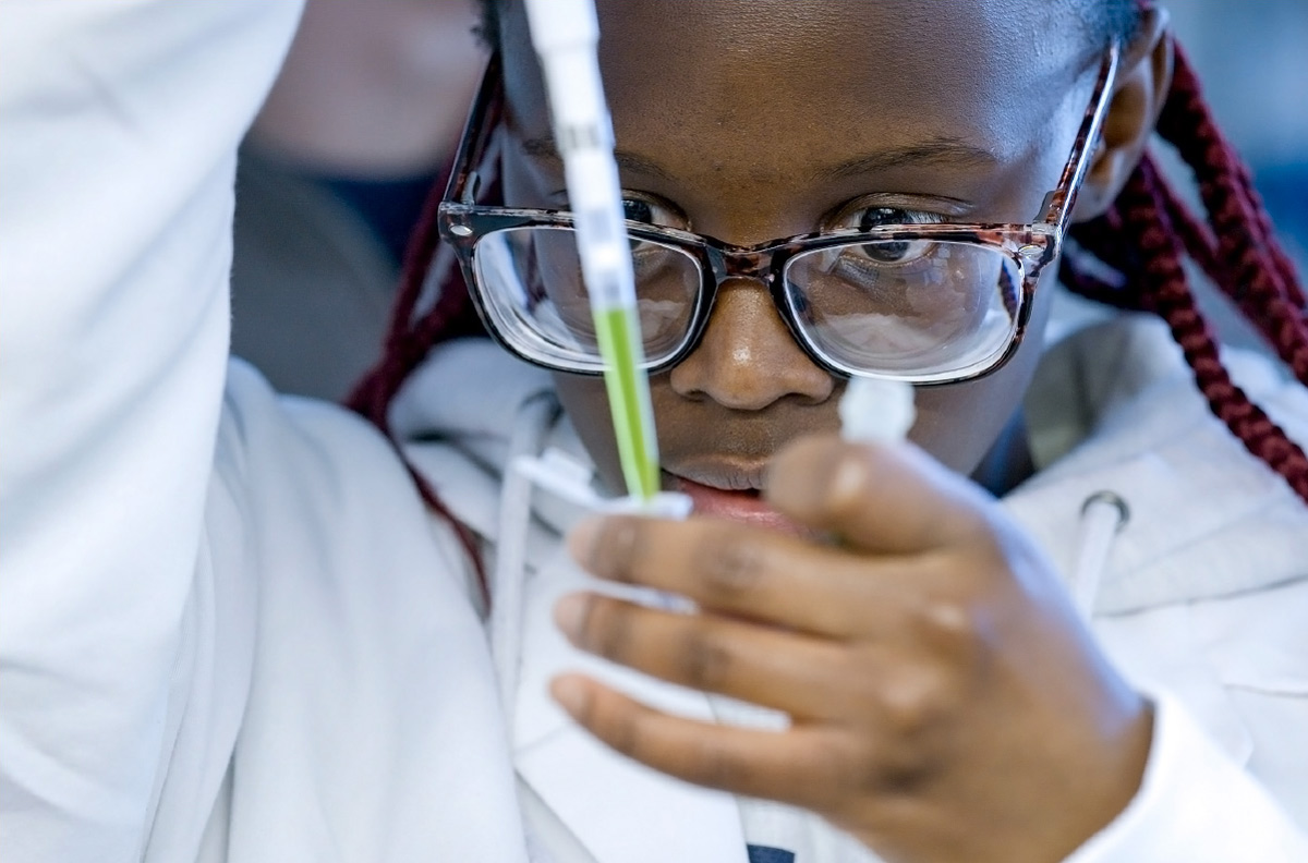 A close-up of a young girl wearing glasses and a lab coat as she intently uses a pipette to measure green liquid in a science laboratory.