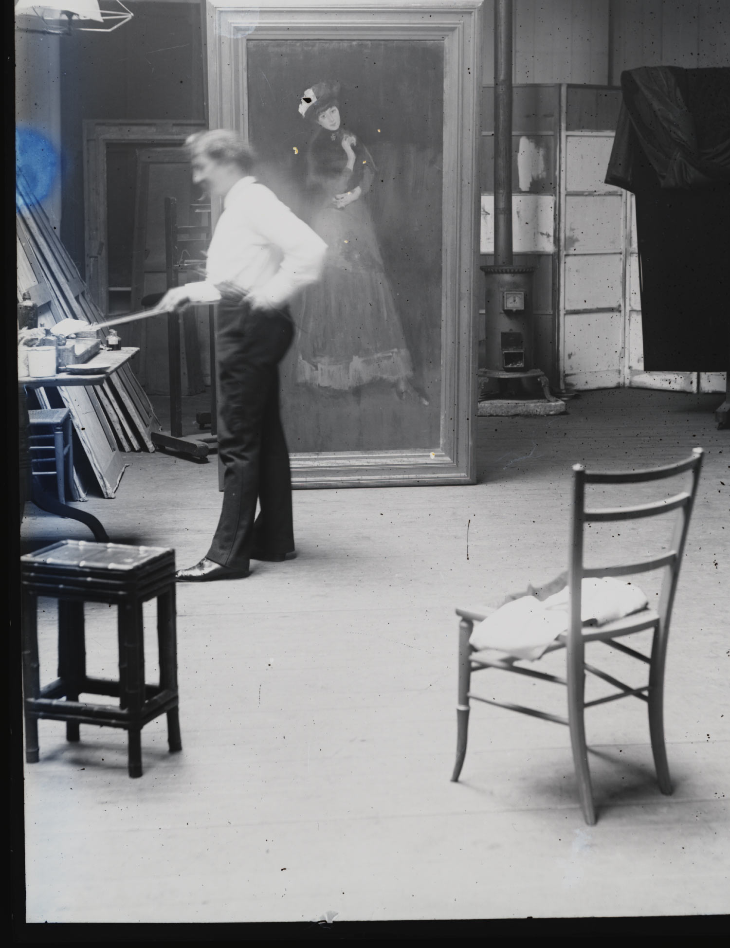 A black-and-white photo of a man painting a full-length portrait of a woman inside a spacious, rustic art studio with chairs and tables.