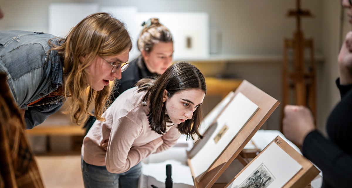 Two students lean forward in an art archive gallery to closely study matted black-and-white prints displayed on wooden tabletops.