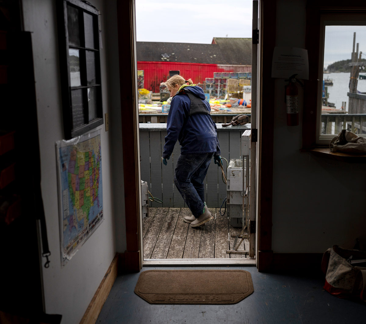 Seen through a dark doorway, Cait Cleaver walks across a wooden deck carrying plastic totes. A red building and stacked fishing traps are visible in the background.