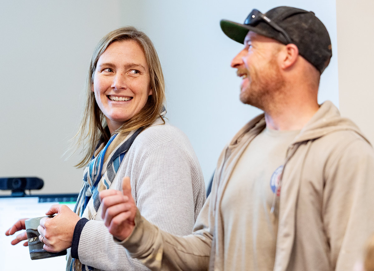 Cait Cleaver smiles while holding a mug next to her husband, Kris Koerber, who wears a baseball cap and speaks during a discussion.