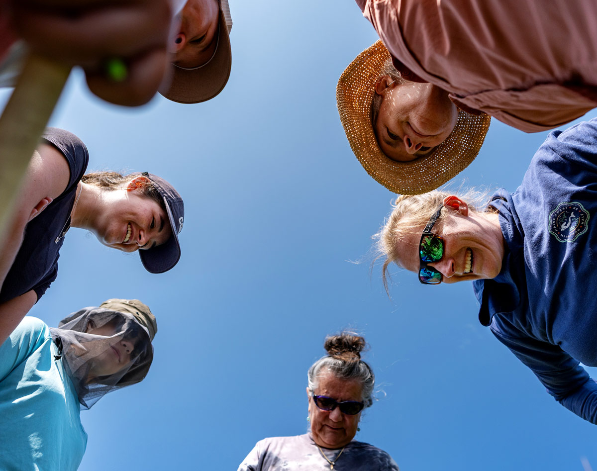 Low-angle view looking up at a circle of researchers and students, including Cait Cleaver, leaning inward against a clear blue sky.
