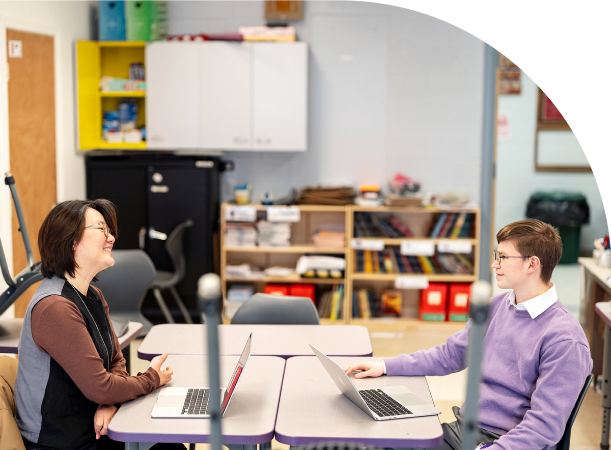 Assistant Professor Andie Wang and Avery Munns sit at individual classroom desks facing each other with open laptops, engaged in a conversation.