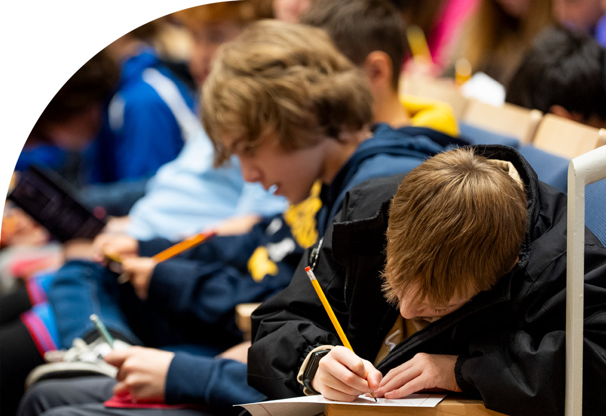 A row of students in auditorium seating lean forward, focused on writing on paper with yellow pencils.