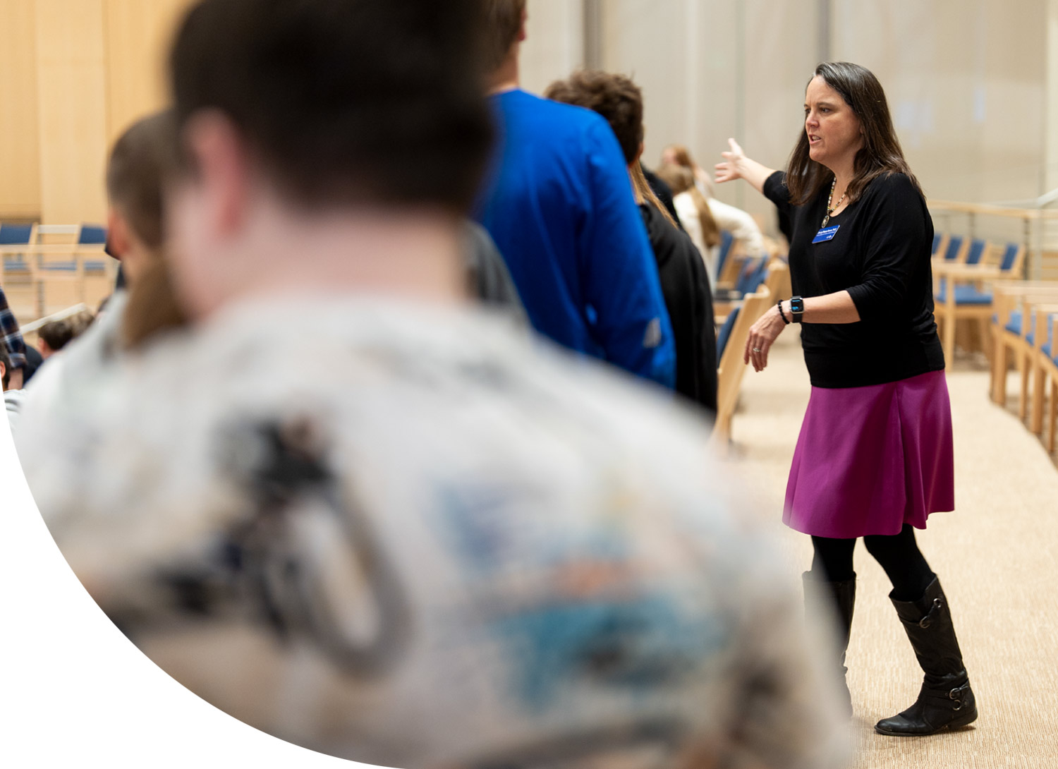 Sherry Pineau Brown stands in an auditorium, wearing a black shirt and a magenta skirt while gesturing with an outstretched arm toward a blurred group of people.