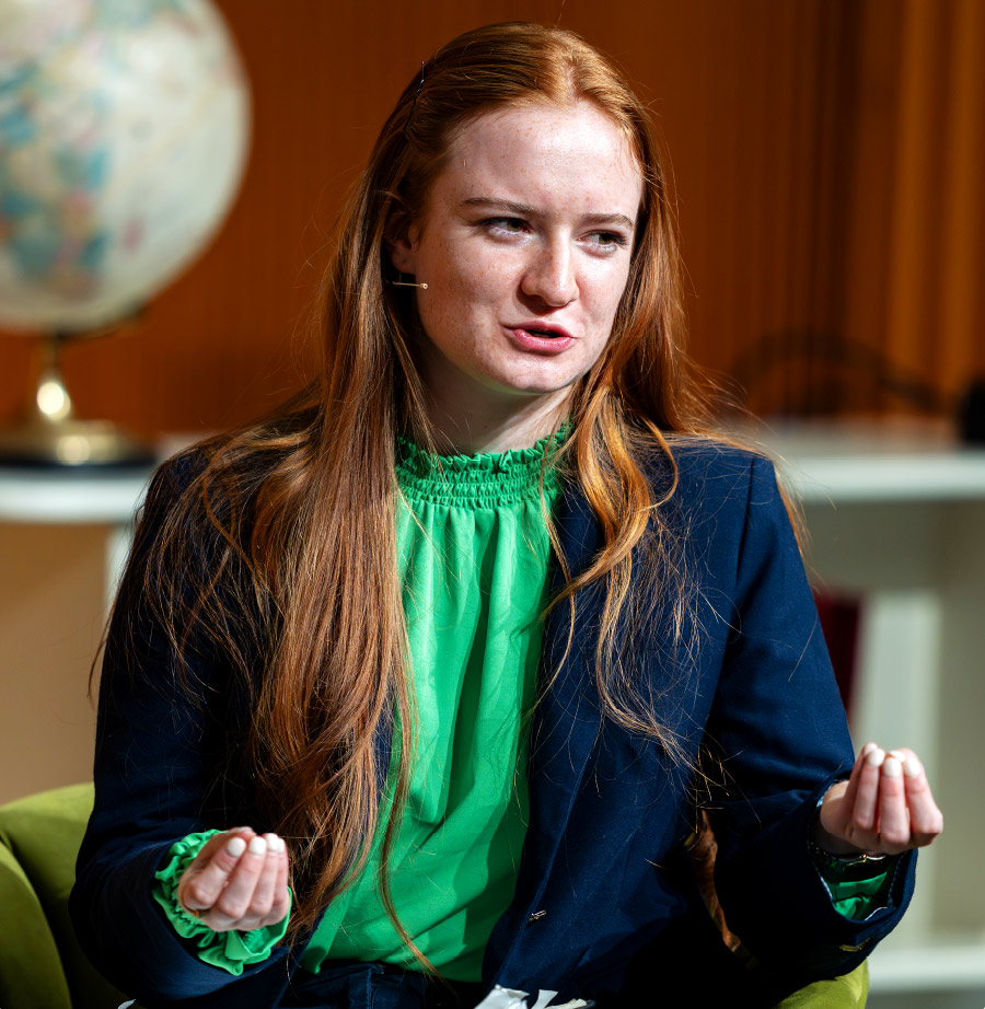 A young woman with red hair gestures while speaking at a university event.