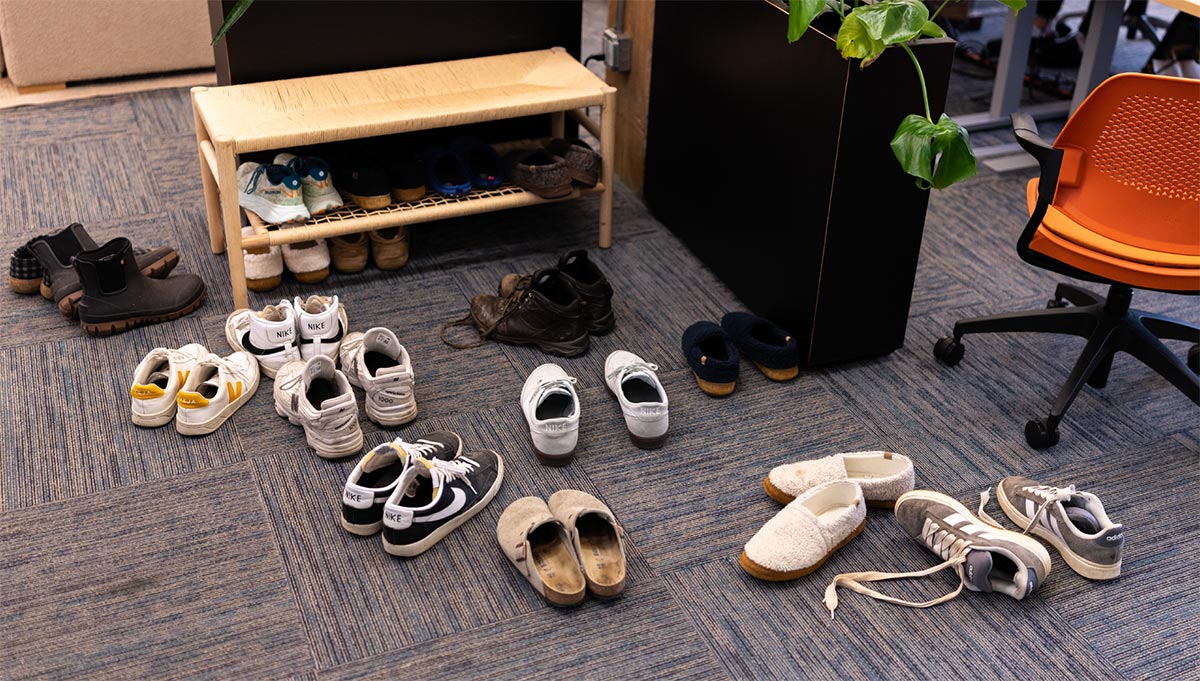 Numerous pairs of shoes and slippers discarded on the office carpet near a wooden rack.